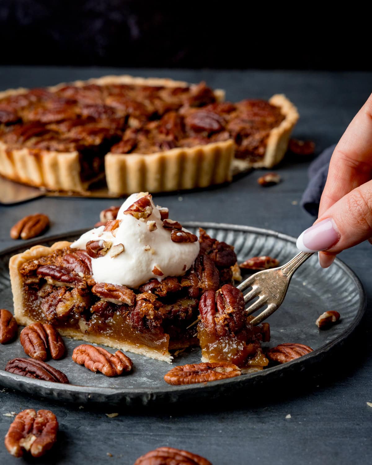 A slice of pecan tart topped with whipped cream on a plate, with a fork held by a hand. A whole pecan pie is visible in the background.