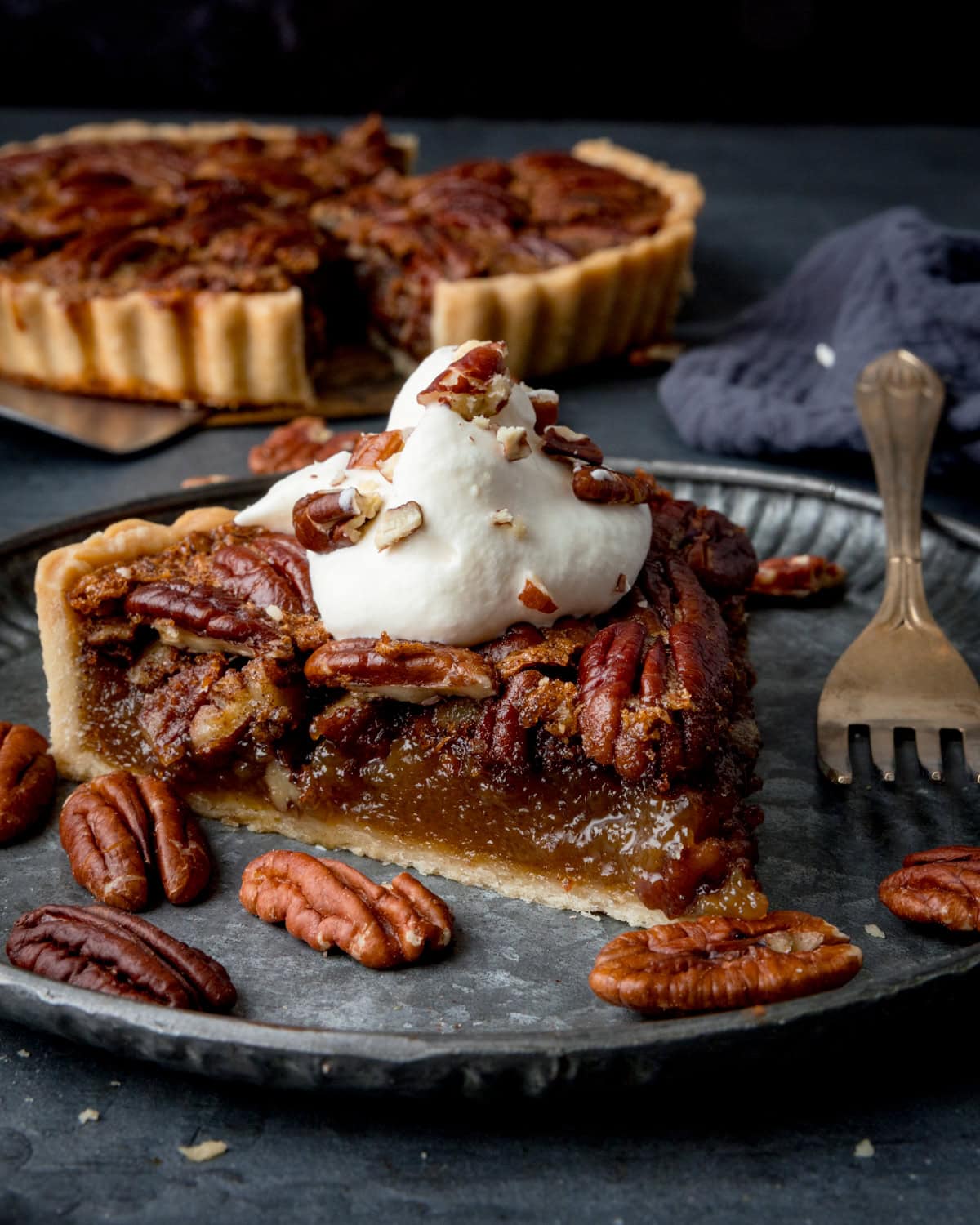 A slice of pecan pie topped with whipped cream on a metal plate, with a whole tart in the background.