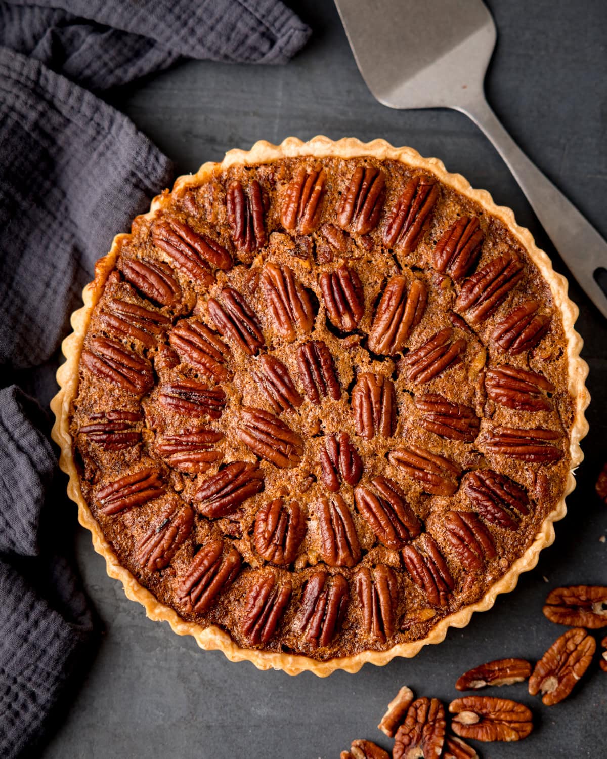 Overhead picture of a pecan pie in a fluted pastry case, with a metal cake slice and loose pecans on a dark surface.