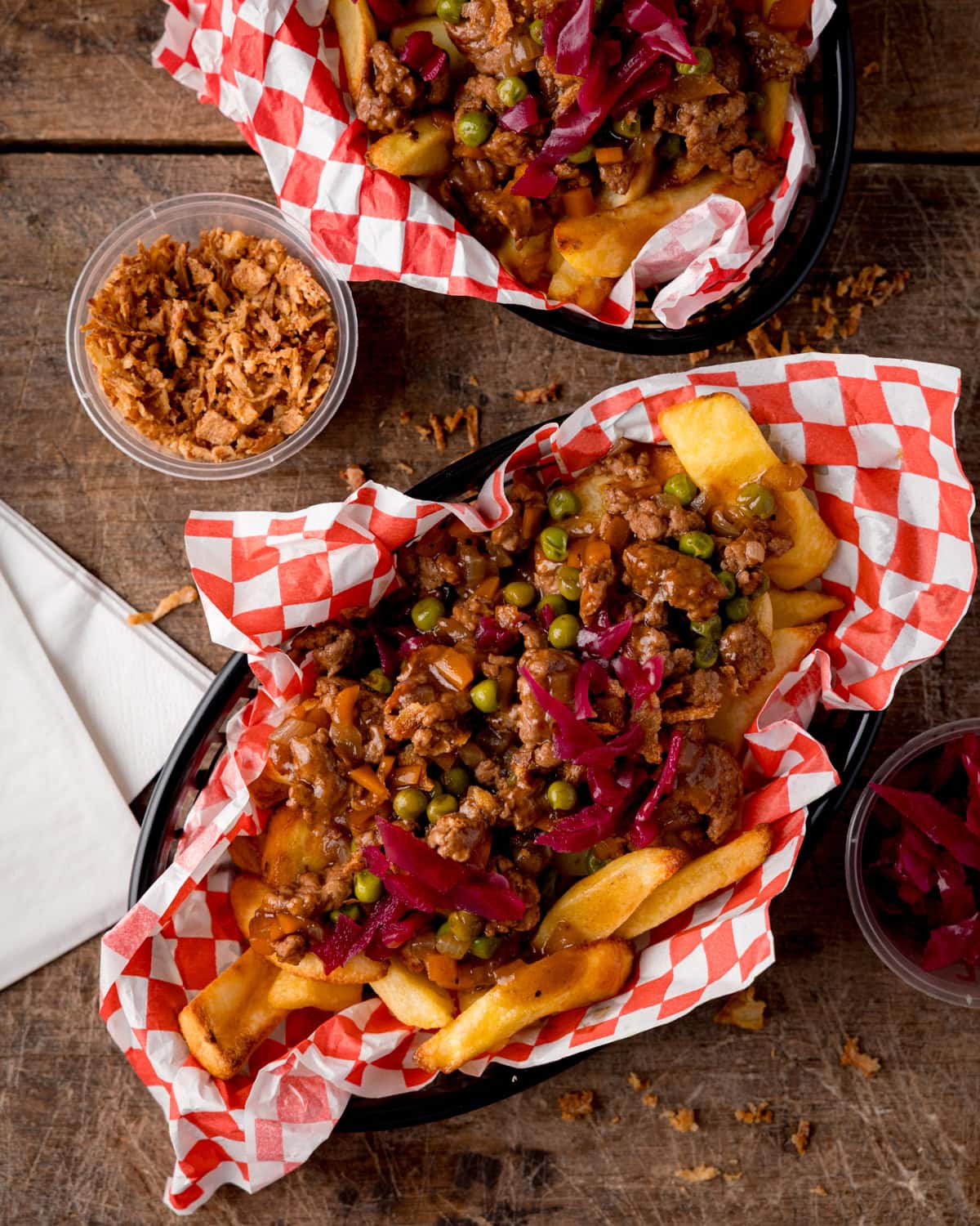 Two baskets of shepherd’s pie chips topped with mince, peas, and pickled red cabbage, served with sides of crispy onions and cabbage.