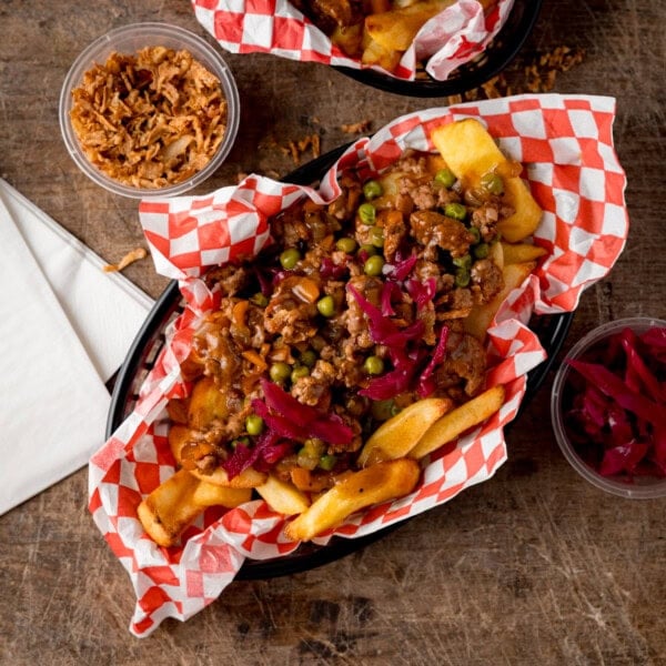 Shepherd’s pie chips topped with lamb mince, peas, and pickled red cabbage in a checkered paper basket, with side containers of crispy onions and extra cabbage.